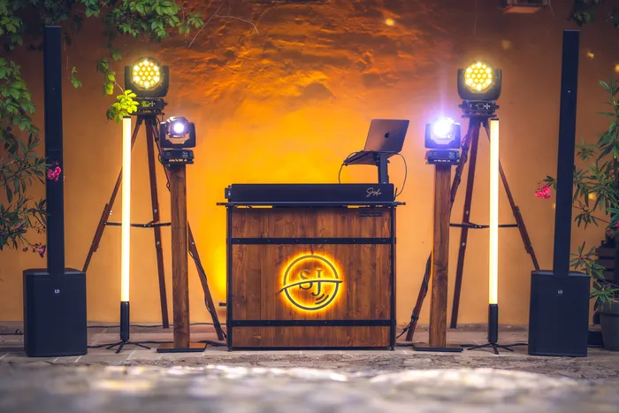 DJ-Pult mit SJ-Logo, Moving-Heads und Lautsprechern vor oranger Wand
