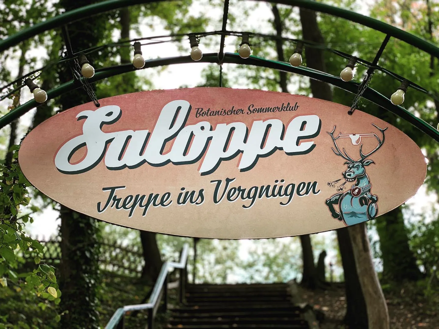 Ovales Eingangsschild mit Lichterkette zwischen den Bäumen
